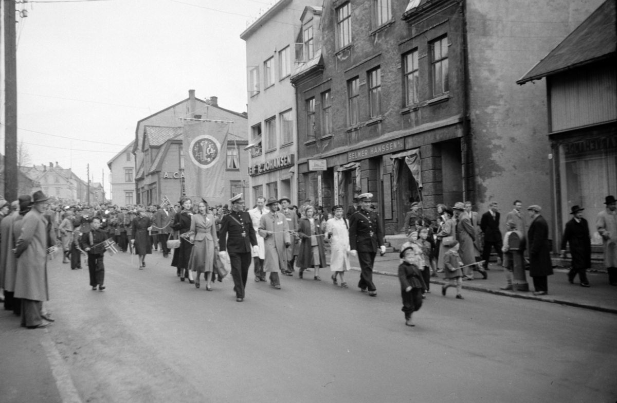 17. maitoget marsjerer ned Strandgata(?), med to politimenn i front. I bakgrunnen ser vi blant annet butikkene til A. Carlsen,  Leif Johansen og Selmer Hanssen.