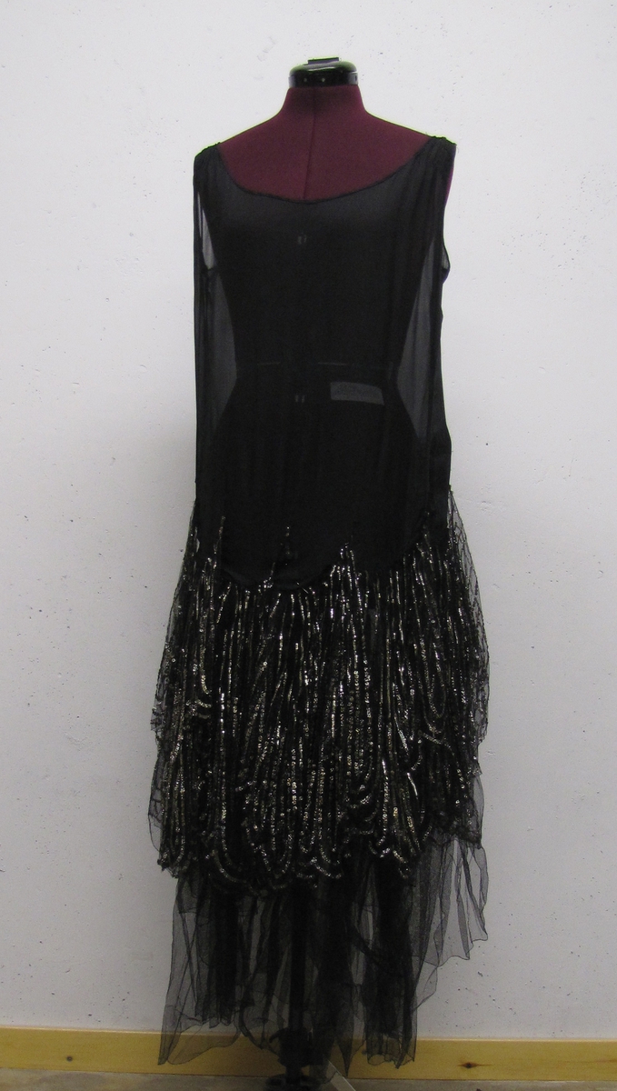 Ärmlös klänning, avskuren vid höften. Kjolen består av tyll, broderad med silverpaljetter. Underkjol av svart tyll.

Klänningen är från 1920-talet.