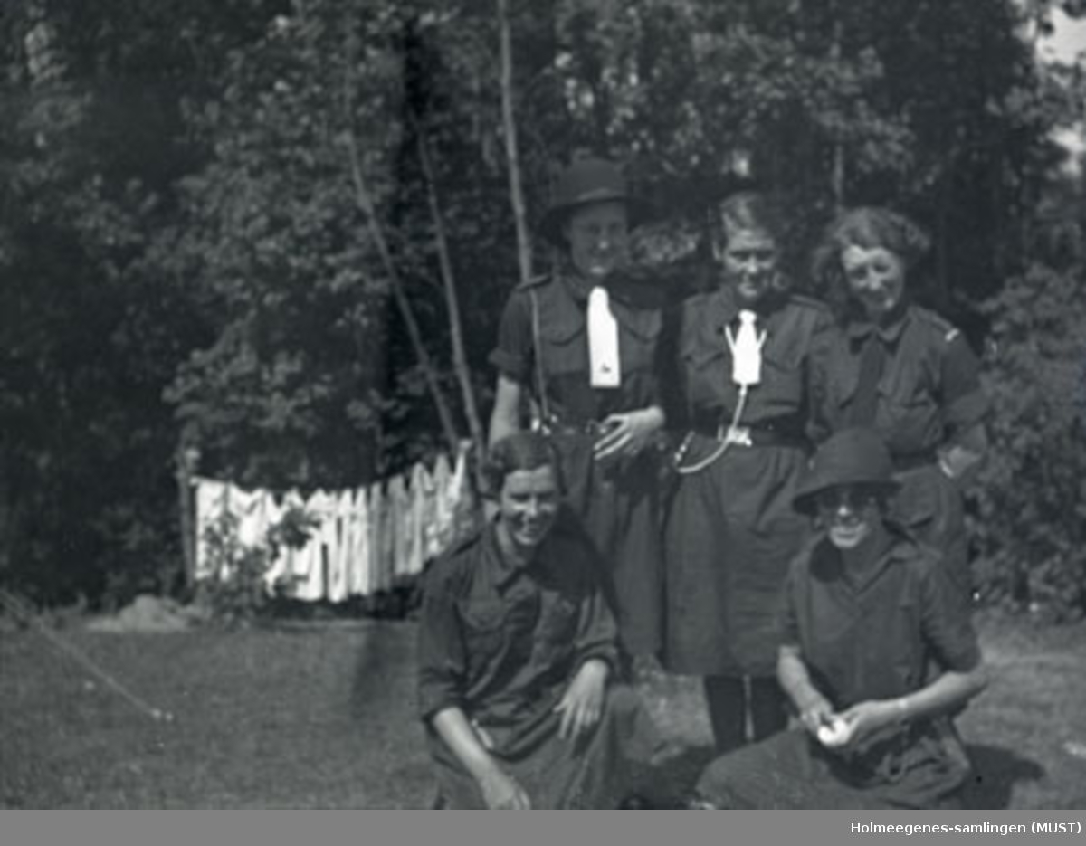 Fem speidere (jenter) med speiderdrakt poserer for fotografen. I bakgrunnen ses et skogholt med klesvask hengt til tørk.