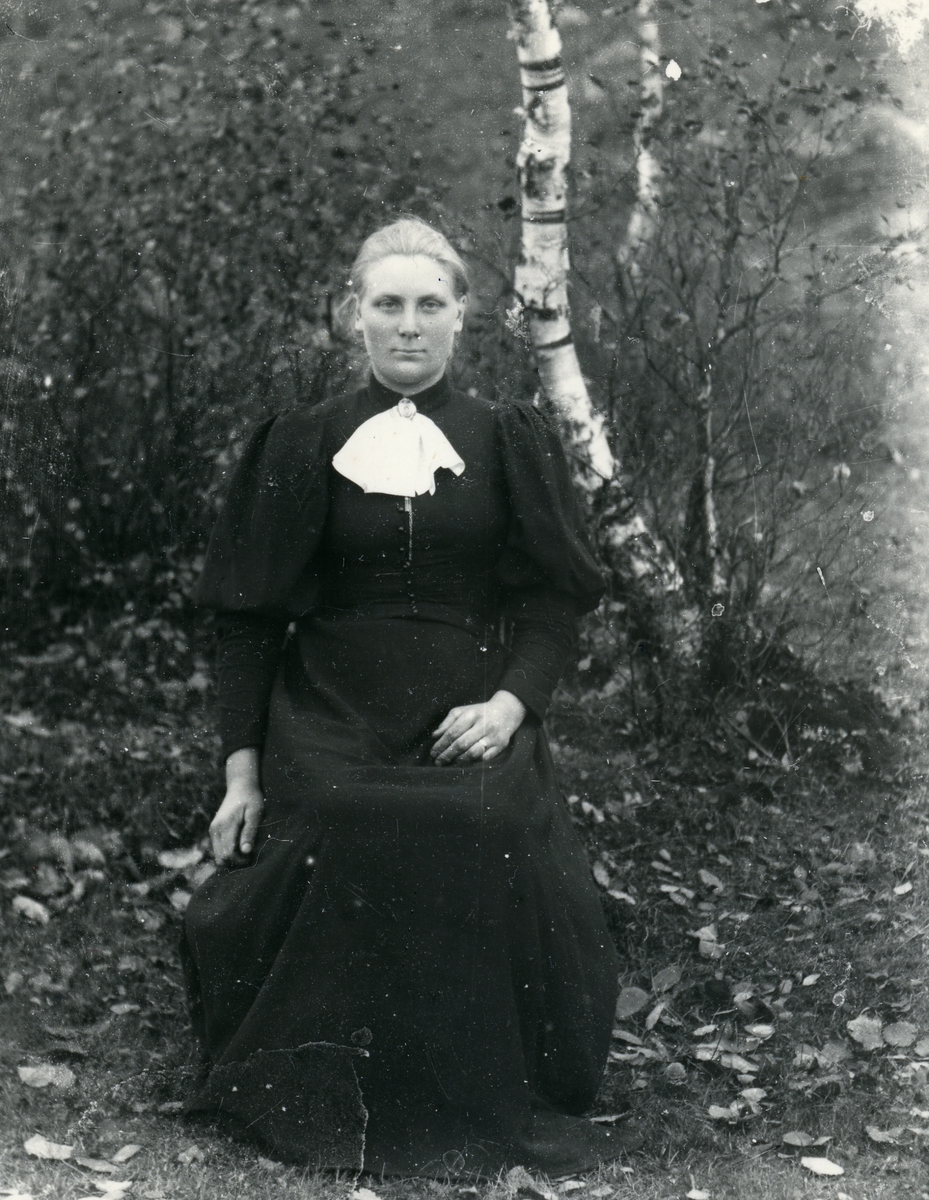 Kvinne sittende på stol, kledd i mørk kjole og kalvekryss, med busker og bjørketrær i bakgrunnen