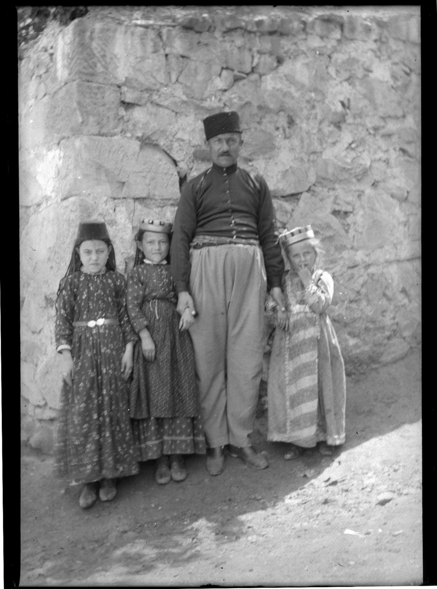 'Grupp med 1 man och 3 barn, flickor stående framför vägg. Kläder. Klädsel. ::  :: Ingår i serie med fotonr. 5280:1-17. Se även fotonr. 5269-5292 med bilder från Stuxbergs resa till Kaukasien.'
