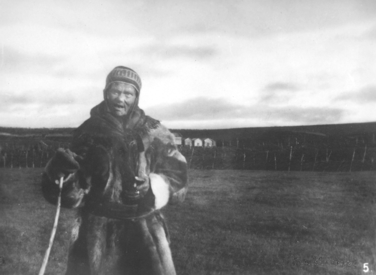 Bilde nr. 5 i serien '10 amatørbilleder fra lappernes hjem og liv i Finnmark', se FB 93164-001. 'Lappekone, i Kautokeino paa vei til kirke.'  Kvinnen er kledt i pesk, lue, votter og skjerf. I handa har hun en kjepp. Det er ikke snø.
