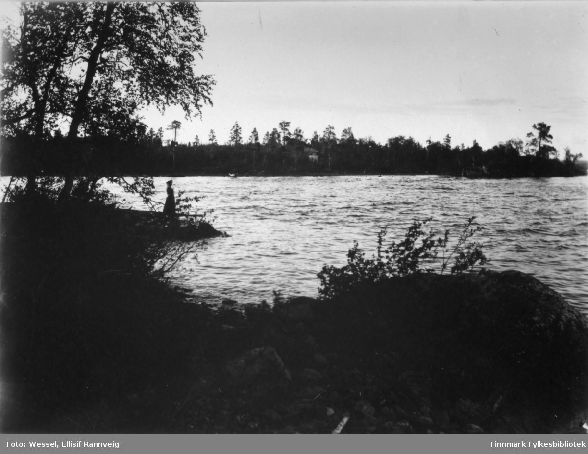 Et landskapsbilde fotografert fra Pasvik. Bak bildet står teksten 'Fra Mennikafoss i Pasvik mellom fossens to fald.' Ved stranden står en kvinne. På andre siden elvaen ligg det en hytte.
