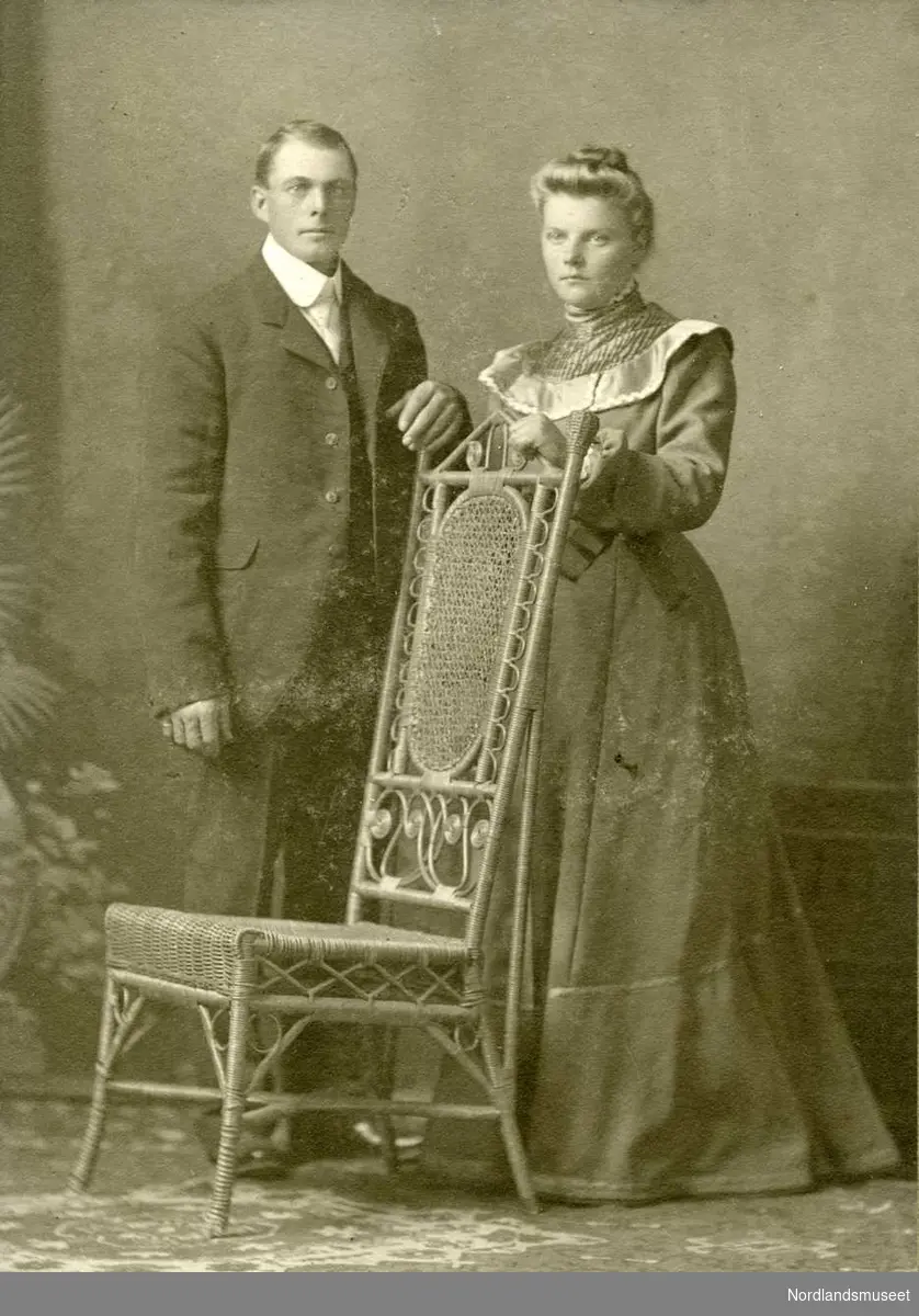 Atelierportrett av ungt pent kledt par stående ved flettet stol med høye ryggen.
Helfigur.