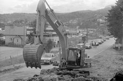 Gravemaskin. Biler i kø på veien i bakgrunnen.