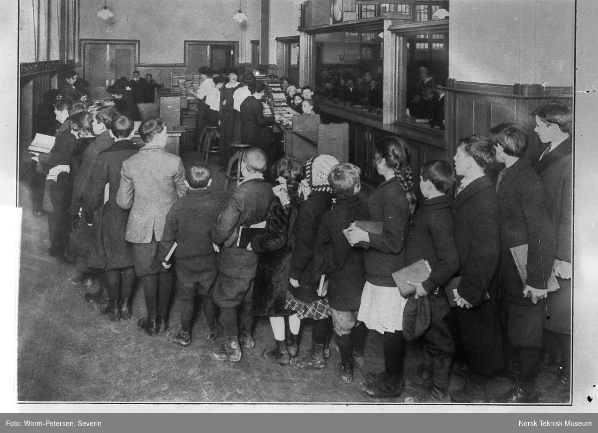 Biblioteksinteriør med kø av barn foran skranke. "Waiting their turn at the Brownsville Childrens Branch". Avfotografert.
