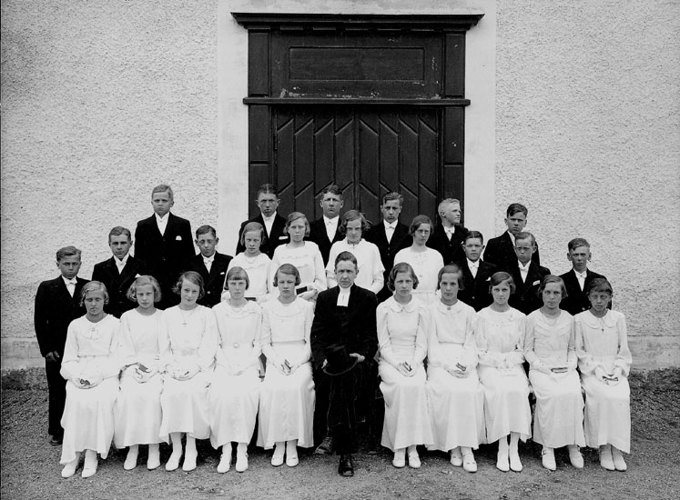 Konfirmander, 14 flickor, 12 pojkar och pastor Eriksson.
Kyrkobyggnad i bakgrunden.