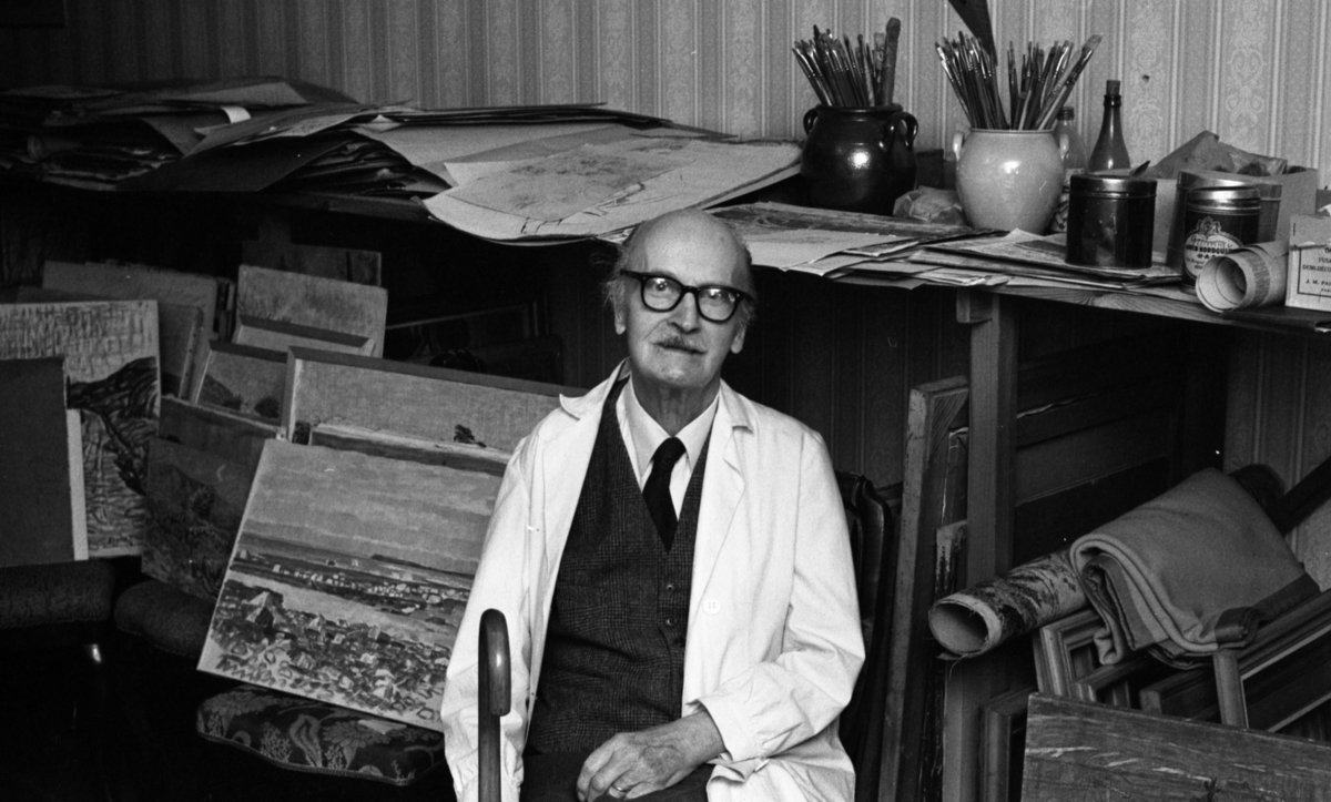 Frans Timén 30 december 1967

Konstnär sitter på en stol i sin atelje. Bakom honom ser man en bänk med krukor och penslar, och tavlor med ramar och utan ramar.