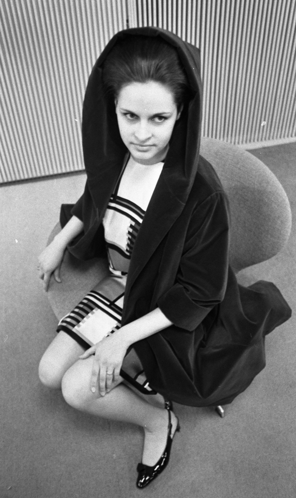 Modebilder 23 april 1966

En mannekäng klädd i svart kappa med huva, kort mönstrad ljus klänning och svarta lågklackade skor sitter på en stol.