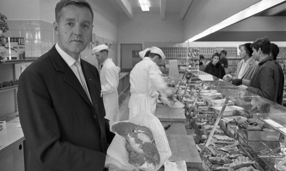Ny Varberga affär, 22 oktober 1965.

Närbild på butiksinnehavaren som håller upp en oxlägg i papper. I bakgrunden betjänar charkpersonalen kunder över den upphöjda glasdisken. De är klädda i vita rockar och vita båtmössor. Under den bär herrana slips och vit skjorta.