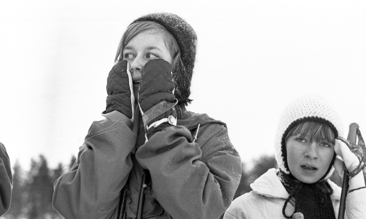 Hästnäs, 20 januari 1966

På bilden syns det ansiktena av två flickor. I bildens förgrund centralt syns det en flicka som är klädd i vinterkläder. Flickan har vantar på sina händer som hon håller vid sina kinder, handflatorna vända mot kinder. Den andra flickan som syns åt höger i förgrunden har en ljus stickat vintermössa på sitt huvud.