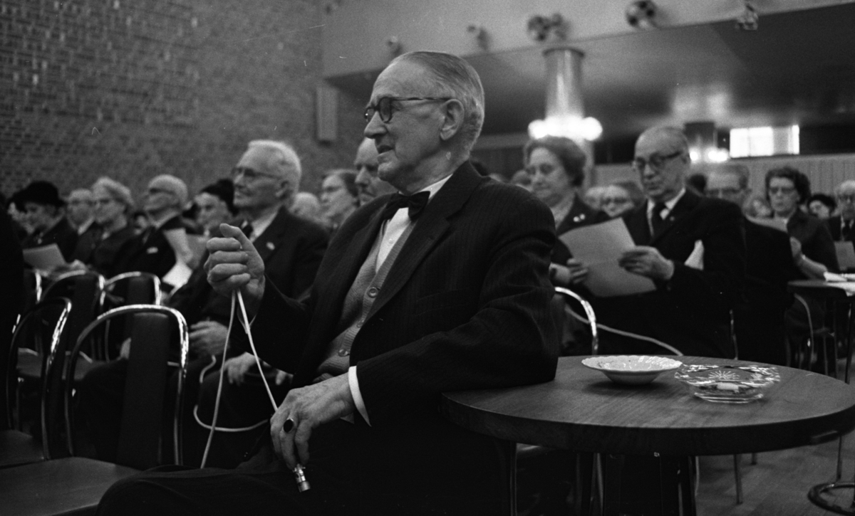 Pensionärspop 2 mars 1966

En äldre herre klädd i kostym, skjorta, kofta och fluga sitter i förgrunden och håller en mikrofon i sina händer. Han stödjer sin vänstra armbåge på ett litet runt bord. På bordet står två askfat. I bakgrunden syns ytterligare damer och herrar som sitter och sjunger.