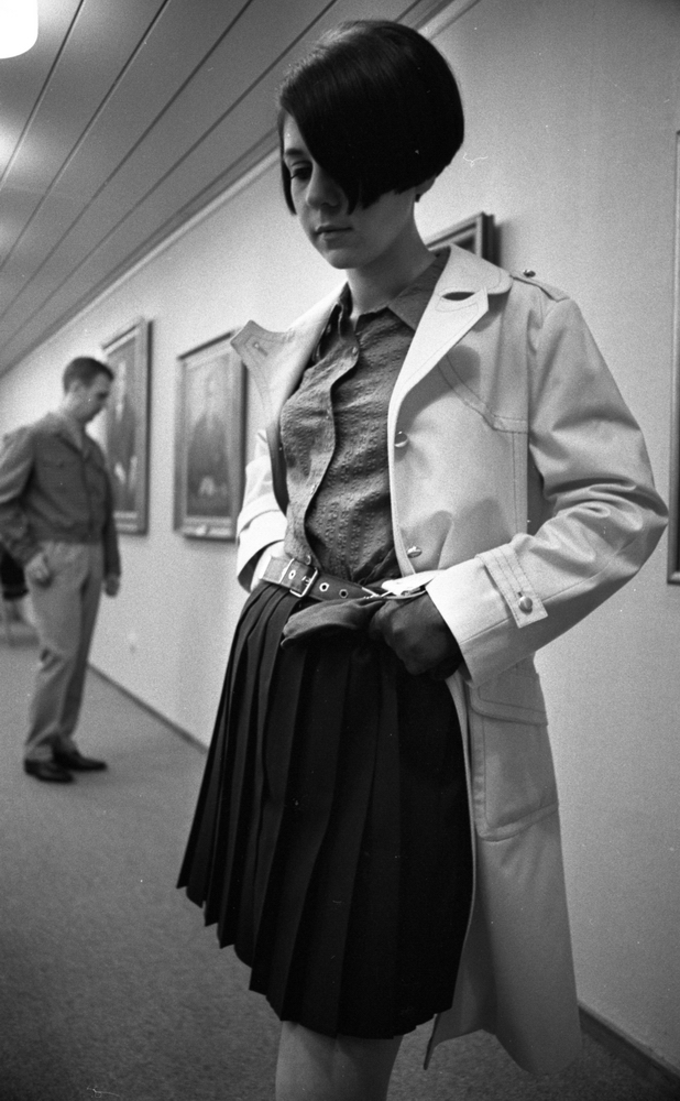 Mannekänguppvisning, Avtack kvinna 18 april 1967

En ung kvinna som är mannekäng poserar i vit kappa, ljus blus, svart veckad kjol och handskar. En man står i bakgrunden klädd i ljus jacka, ljus skjorta, ljusa byxor och mörka skor. Tavlor hänger på väggen. Mannen står och betraktar en av dem.