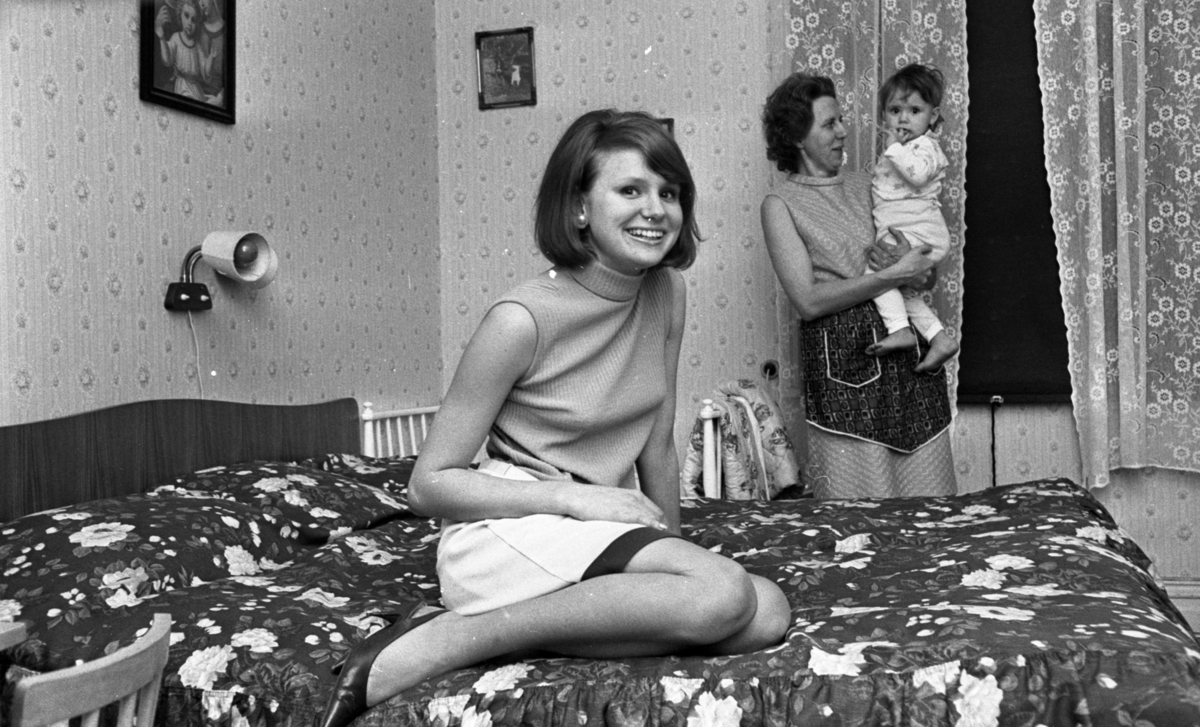 Miss Örebro 14 november 1966

En ung kvinna som blivit vald till Miss Örebro sitter på en säng med blommigt överkast. Kvinnan är klädd i en ljus, ärmlös blus, vit kjol med svart kant nedtill samt ett vit skärp runt midjan. Hon har svarta högklackade skor på fötterna. I öronen har hon örhängen i form av stora vita pärlor Hennes barn har på sig en vit pyjamas med figurer på. Barnet befinner sig i famnen på en kvinna som troligtvis är dess mormor. Den äldre kvinnan är klädd i vit, ärmlös klänning med förkläde över.