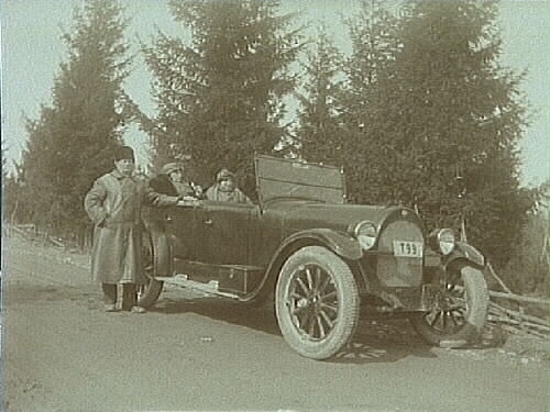 Jenny med sin mor i baksätet på bilen och herr Larsson står brevid. 
Bilen är en Oldsmobile modell 30 från 1923-24 med en 42 hästkrafters sexcylindrig motor. Den registrerades den 2:e april 1924 på fröken Jenny Larsson, Örebro, och fick registreringsnummer T99. År 1930 ägdes den fortfarande av Jenny Larsson.