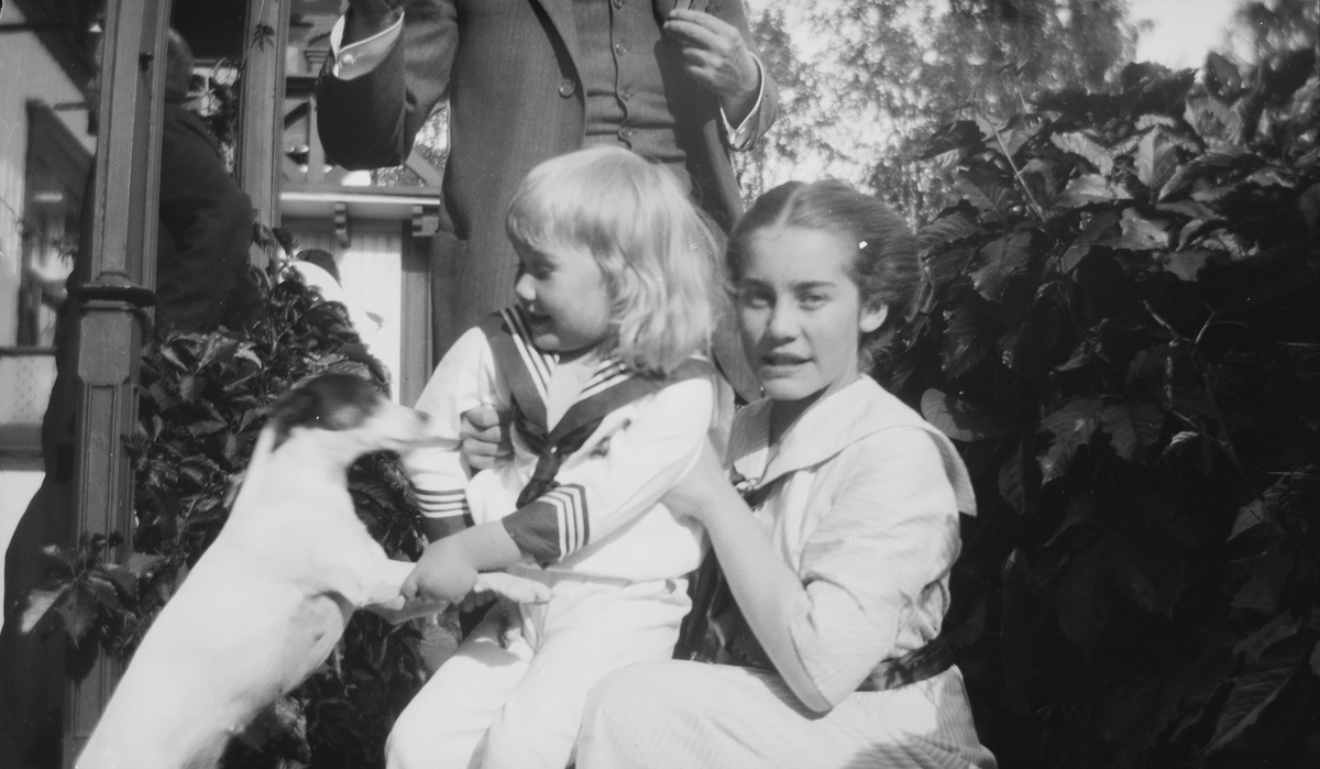 Iacob Ihlen Mathiesen kledd i lys matrosdress leker med en liten hund. Celina Marie Mathiesen sitter på huk og holder barnet. I bakgrunnen står en mann i dress, og en annen lener seg mot glenderet.
