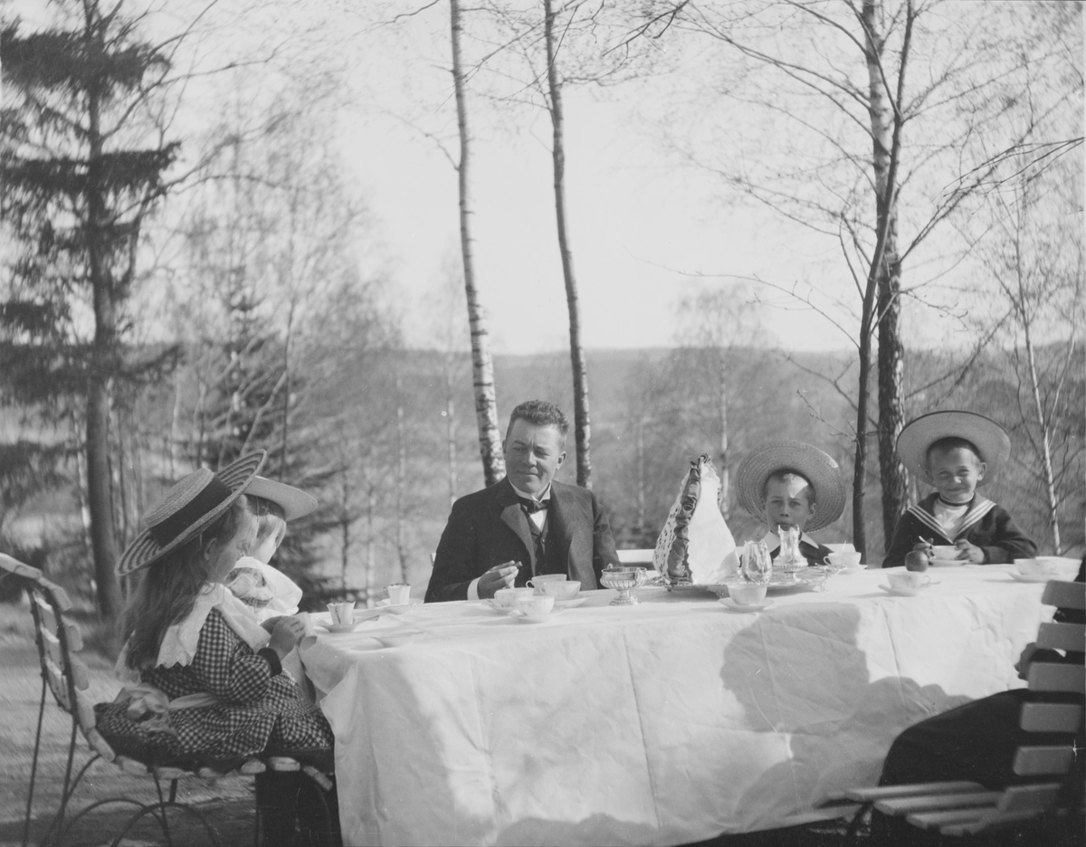 Statsråd Christian Pierre Mathiesen med Christian Pierre f. 1897 og Erich Monsen til høyre i bildet, og Ise (Louise) og Iacob til venstre i bildet ved et bord med hvit duk med oppdekking til kaffe og/eller teselskap i hagen. Vi ser en bit av en voksen person til, sannsynligvis en kvinne.
