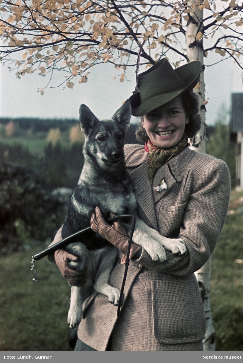 Kvinna klädd i tweed med en hund  i famnen och en läderpiska i sin hand.