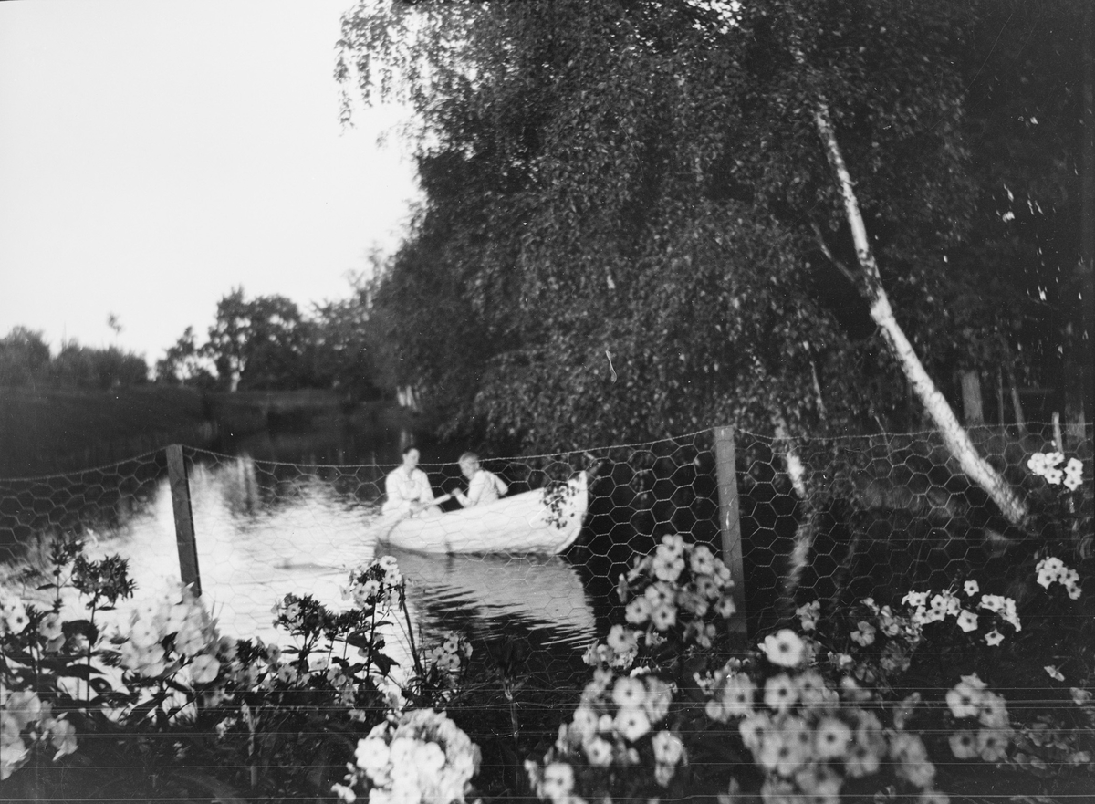 En gutt er ute å ror på karpedammen også kjent som "kanalen". Dammen ligger nederst i terrassehagen på Linderud Gård og går langs med "Lindehallen". En kvinne sitter bak i båten.