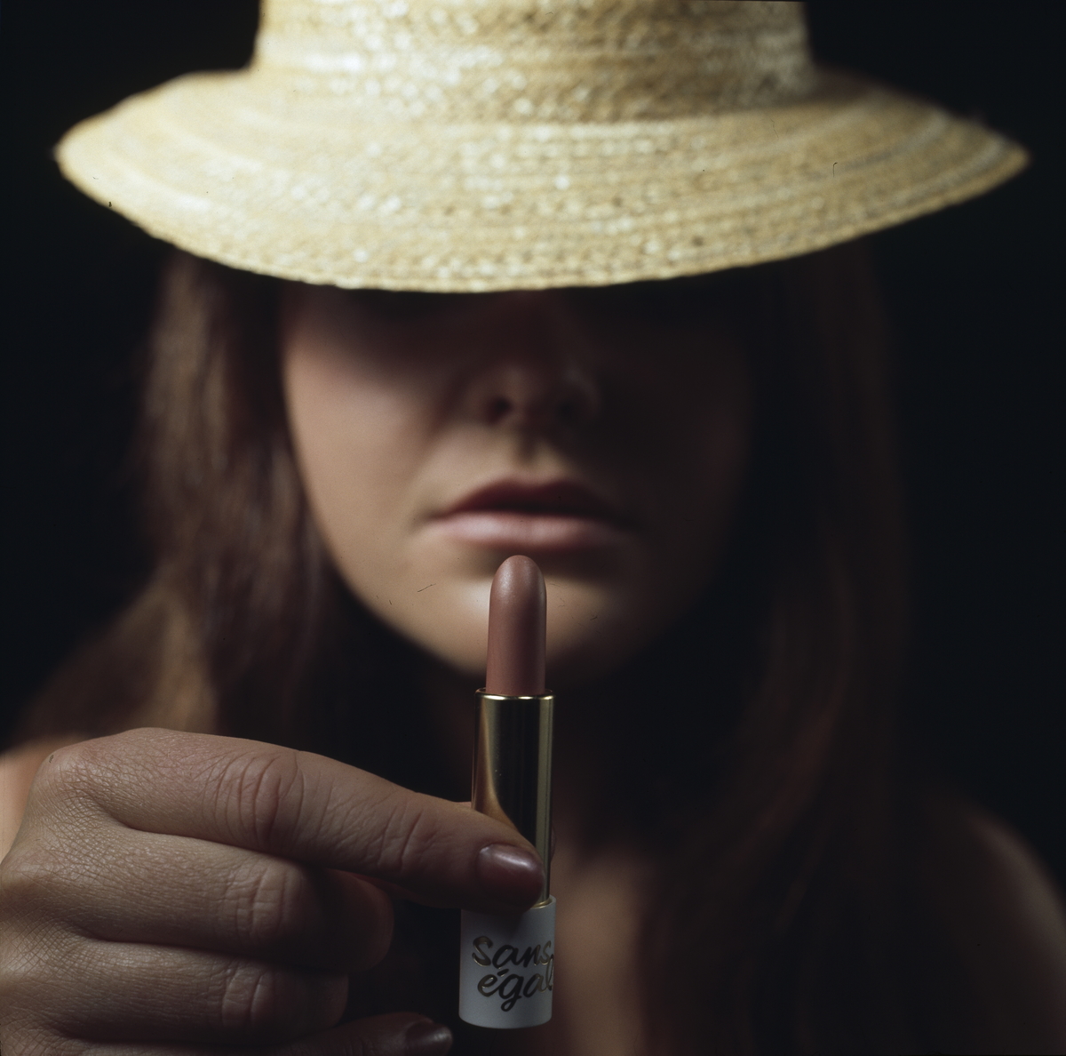Reklamefoto for leppestift. Nærbilde av kvinne med hatt som holder en leppestift opp foran ansiktet sitt..