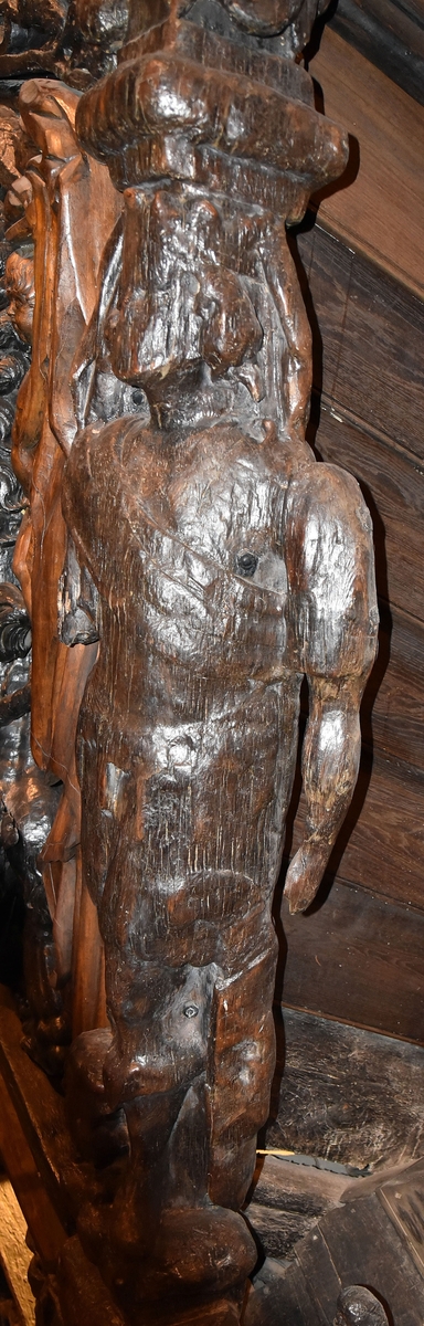 Skulptur i form av en hög, rikt snidad pelare eller stolpe, med kvadratisk genomskärning.

Tre av pelarens sidor är skulpterade medan den fjärde utgör anliggningsyta, försedd med ett antal olikformade urtag. De skulpterade sidorna är horisontellt indelade i tre eller fyra sektioner eller nivåer, avgränsade från varandra av profilerade och snedställda vulster. Nedtill avslutas skulpturen av en ornerad och på mitten insvängd formation.



Ovanför denna nedre avslutning är pelaren utformad som en reslig mansfigur, vänd mot vänster, sedd från betraktaren. Mansfiguren, som täcker tre av skulpturens fyra sidor, är iförd en romersk rustning med en bred fältbindel draperad från höger axel snett över bröstet. På huvudet med det lockiga och utslagna håret sitter en krona av öppen typ, med liljeliknande ornament. Figurens vänstra arm är böjd, medan den högra armen är sträckt utmed sidan. Handen till den högra armen saknas, men har hållit en rikt profilerad sköld, med mycket slitna och vaga konturer av en på bakbenen mot vänster gående grip med stora vingar. Skölden är snidad i ett med figuren, men med kraftiga underskärningar. Vid figurens fötter skymtar ett djur av något slag. Bakom mannens huvud tycks ett snidat textilstycke, kanske del till ett draperi, vara anbragt.



Ovanför den romerska krigaren eller riddaren har pelaren formen av en herm utan armar med långt, krusigt hår och mustaschprytt ansikte. Liksom mansfiguren täcker även hermen tre sidor av pelaren. Huvudet är vridet mot vänster. Överkroppen är bar så när som på ett flikigt plagg som täcker axlar och övre delen av bröstet. Från huvudet och runt midjan är ett tygstycke draperat. Hermens nederdel är formad till en stor maskaron med uppspärrad mun och utsträckt tunga. Maskaronen övergår upptill och utmed sidorna i yppiga bladformationer.



Pelaren avslutas upptill av ett knoppliknande ornament. Ornamentet är ornerat med akantusbladliknande bildningar samt fyra byglar som möts på toppen, där vissa spår antyder att det ursprungligen suttit ett klot eller dylikt, men som fallit bort. Eventuellt kan ornamentet utgöra en huvudbonad till hermen.

Skulpturen är kraftigt sliten.



Text in English: Sculpture in the shape of a richly carved column or pillar.

Three sides of the pillar are carved, while the fourth side comprises the attachment surface and has a number of variously-shaped indentations. The sculptured sides are divided horizontally into three or four sections separated by angled tori.



The lower section of the pillar is carved in the figure of a tall man. The figure is clad in Roman armour and has a field badge (cloak/plaid) draped across his chest at an angle from his right shoulder. On his head he wears an open crown. He has long, curly hair. The right arm is broken off, the left is at his side. The left hand is missing but once held a richly carved shield.

The shield is carved out of the same piece of wood as the figure, but deeply undercut. A carved figure is apparent on the shield. Although the shield is badly worn, it is possible to recognise one of the hind legs of a griffin with large wings. At the man's feet lies an animal of some kind.



Above the Roman warrior or knight, the pillar is carved in the shape of a herm, without arms and with long, curly hair and a moustache. Like the male figure, the herm also covers three sides of the pillar. The head is twisted to the left. The upper part of the body is bare except for a piece of cloth that covers the shoulders and upper chest. The lower part of the herm is carved as a large mask, its mouth open and tongue protruding. The sides and upper parts of the mask are carved as luxuriant leaves.



The upper part of the pillar has a ornament in shape of a knob. The ornament has a ring of acanthus leaves and four spherical loops run vertically from the acanthus leaves and meet at a point at the top. An object, for example a globe appears to have adorned the top of the ornament. This object is missing. The ornament may also be a part of the headgear of the herm.

The sculpture is severely worn.