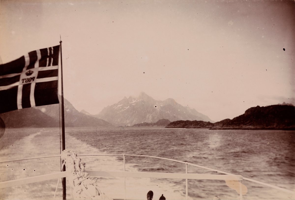 I forgrunnen ser vi akterdekket til postbåten med postens unionsflagg. I bakgrunnen fjord- og fjellandskap.