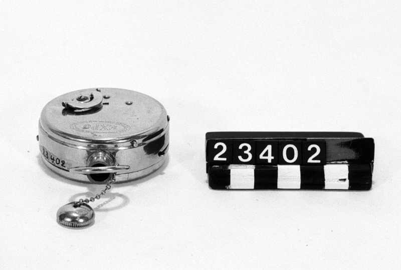 Kamera i form av fickur "Expo watch camera", för rullfilm i specialkassett. Objektivet, en enkel lins, i remontoaröppningen. I kamerans främre del saknas en halvcirkelformad skiva av 2 mm tjock röd fibermassa med tryckt text: EXPO (vitt) pat. througout the world U.S. patent sept. 6. 1904. Denna skiva är på T.M. kopierad 14/4 -62 efter en annan kamera (tillh. Herr Werneström, Sthlm) som dessutom hade ett fodral av ljusbrunt läder.
Tillbehör: Tom filmkassett.