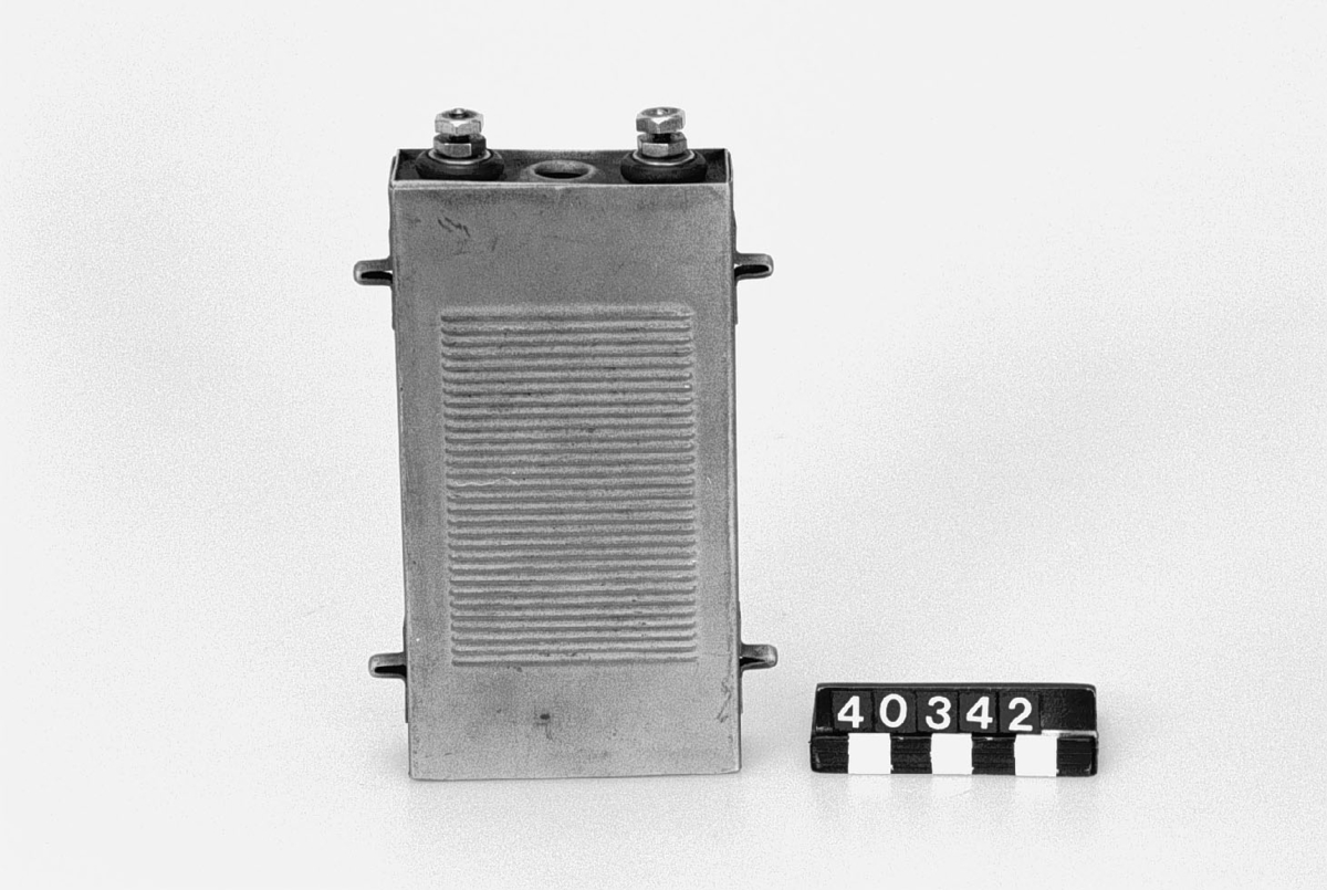 Battericell av plåt, märkt: +, SI, 1. Två anslutningar i toppen. Fyra punktsvetsade "piggar", två på vardera kortsidan.