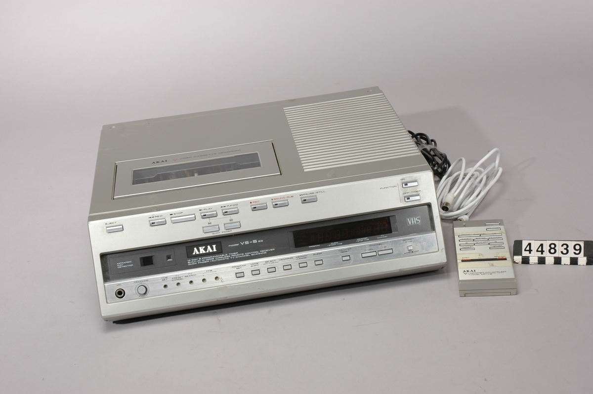 AKAI PAL VHS toppmatad videobandspelare med infraröd fjärrkontroll. Hölje av grå plast, med kontroller och anslutningsmöjlighet för mikrofon på fronten. Anslutning för antenn, apparatsladd samt utgångar för video (RCA) och audio (DIN).  Inbyggd tevemottagare/tuner VHF band I - UHF band IV/V. Inbyggt 24h digitalur med fluorescerande display, insomningsfunktion.Timerinställning för inspelning av högst nio program; fem engångs, två varje-dag och två varje-vecka. 
Fjärrkontroll, bruksanvisning och sladdar medföljer.