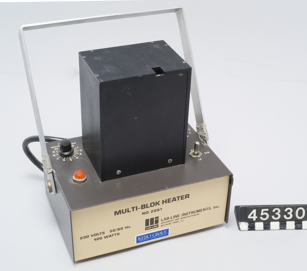 Värmare för preparat i provör. Tillverkare Lab-line Instruments, Inc. modell Multi-blok Heater typ 2091 Till värmaren hör en svart metallbox med plats för sex provrör, det finns en ursparing för termometer i locket.