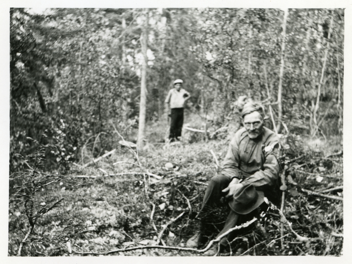 Portrett av en mann sittende i en skog. Mannen har en hatt på fanget og i bakgrunnen kan det ses en annen mann.