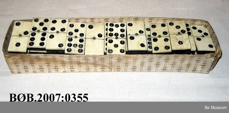 Avlang ask uten lok med dominobrikker
Dominobrikka er sett saman av to delar, underdel av tre og topp av bein. Desse er sett saman med ei messingstift. Det er fresa hol litt ned i beinet. Desse hola er måla. Det er 47 dominobrikker