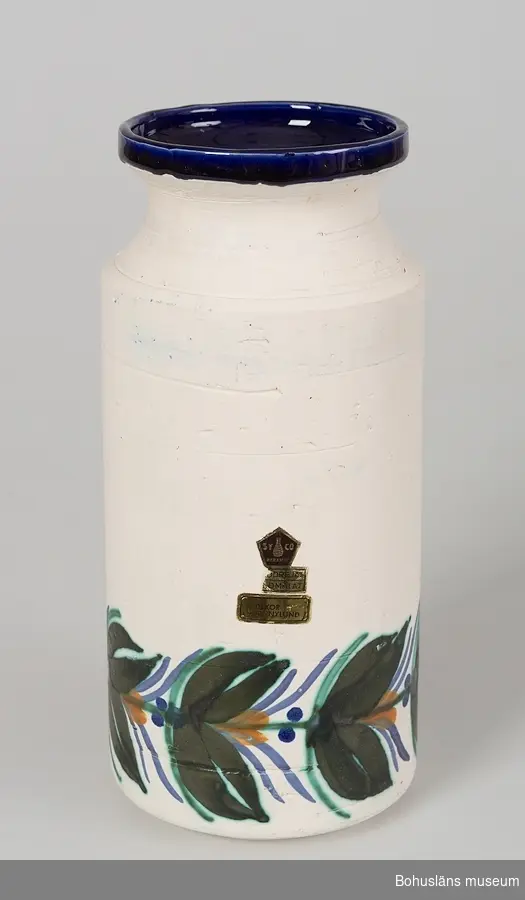 Cylinderformad, konkavt mynningsparti. Vit, matt glasyr. Nedtill runt vasen är den handmålad med stiliserad bård i mörkgrönt, ljusgrönt, blått och orange. 
Invändigt täckt med blank, blå glasyr.
På sidan fastklistrad femkantigt fabriksmärke av papper för "SYCO - keramik" med texten: "Handdrejat, handmålat" samt rektangulärt märke "A.Nylund". Under botten märkt i blått: "AN". 
Av givaren inköpt på Uddevalla Auktionskammare.
