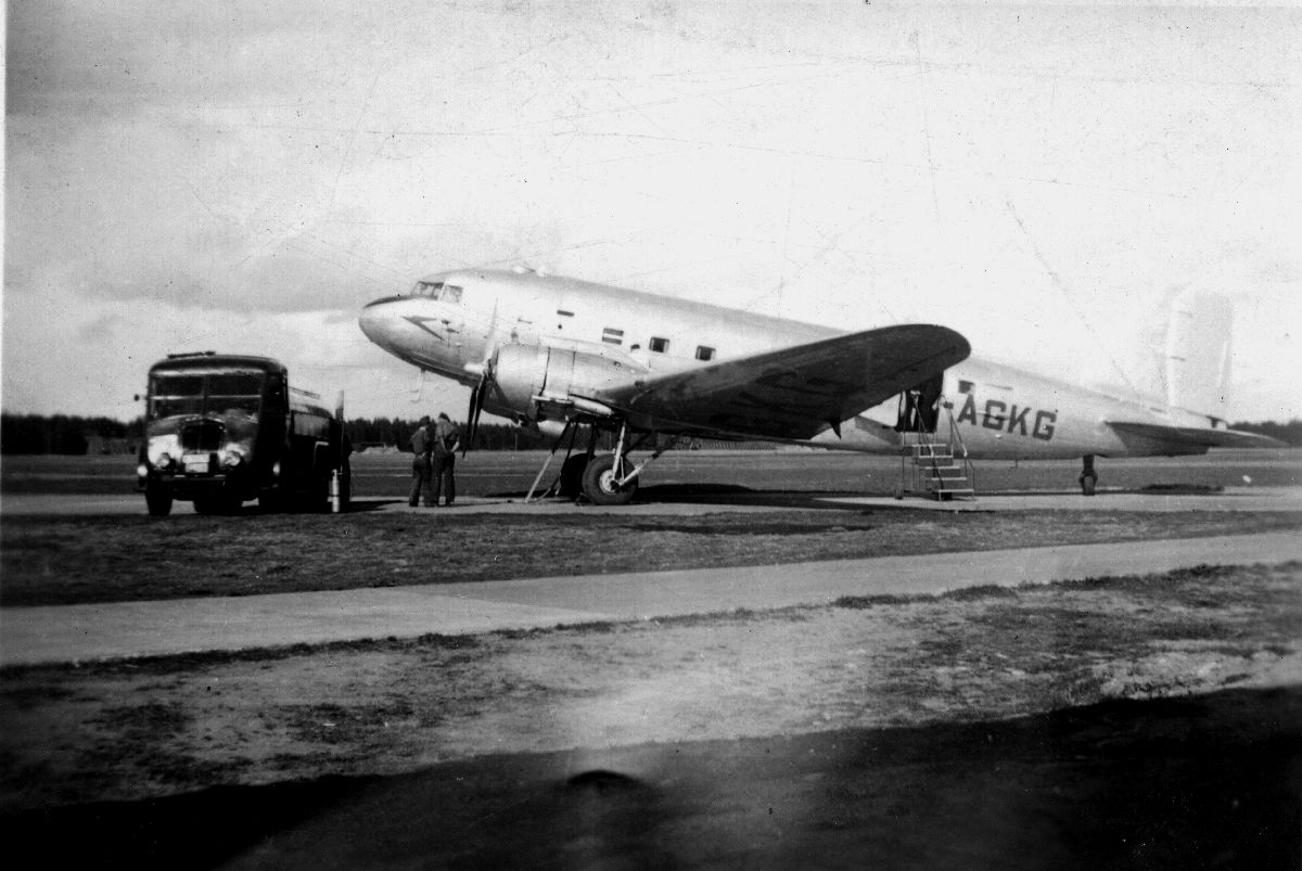 Lufthavn/Flyplass. Et fly, Douglas DC3 Dakota, parkert. En tankbil og to personer fyller drivstoff til neste avgang.