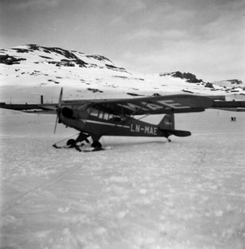 Landskap. Et fly, LN-MAE, Piper Cub fra Widerøe, med ski/hjul understell, står parkert på et islagt vann. I bakgrunn snødekte fjell.
