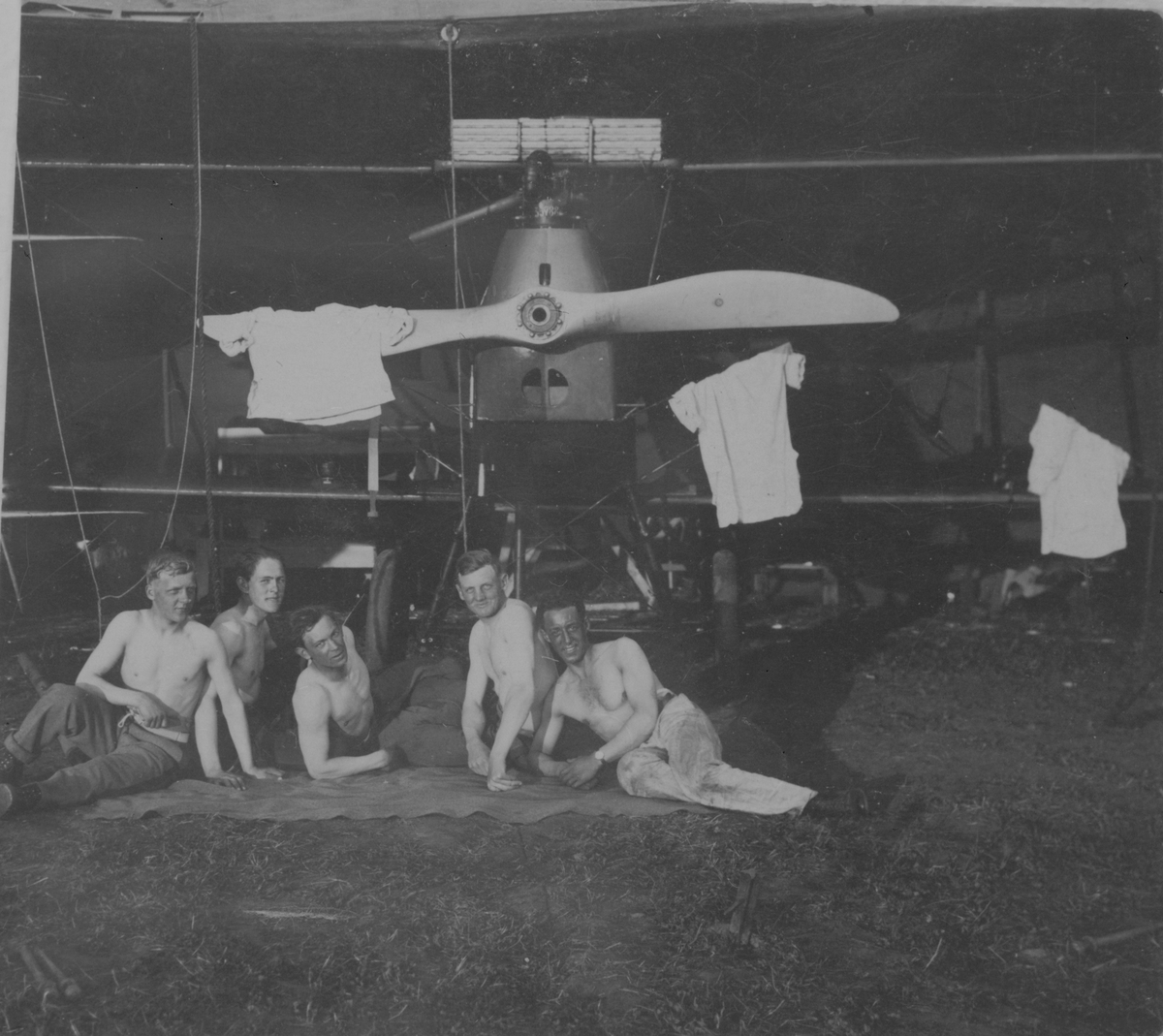 Sommardag. Fem män med bar överkropp sitter framför ett flygplan i en hangar på Hästholmens flygfält, 1923. På flygplanspropellern hänger tröjor.