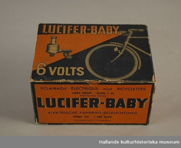 Svart och orange hopnitad pappask för cykelbelysning. På kartongen är framdelen av en cykel avbildad. På insidan av locket står en text skriven med blyerts.