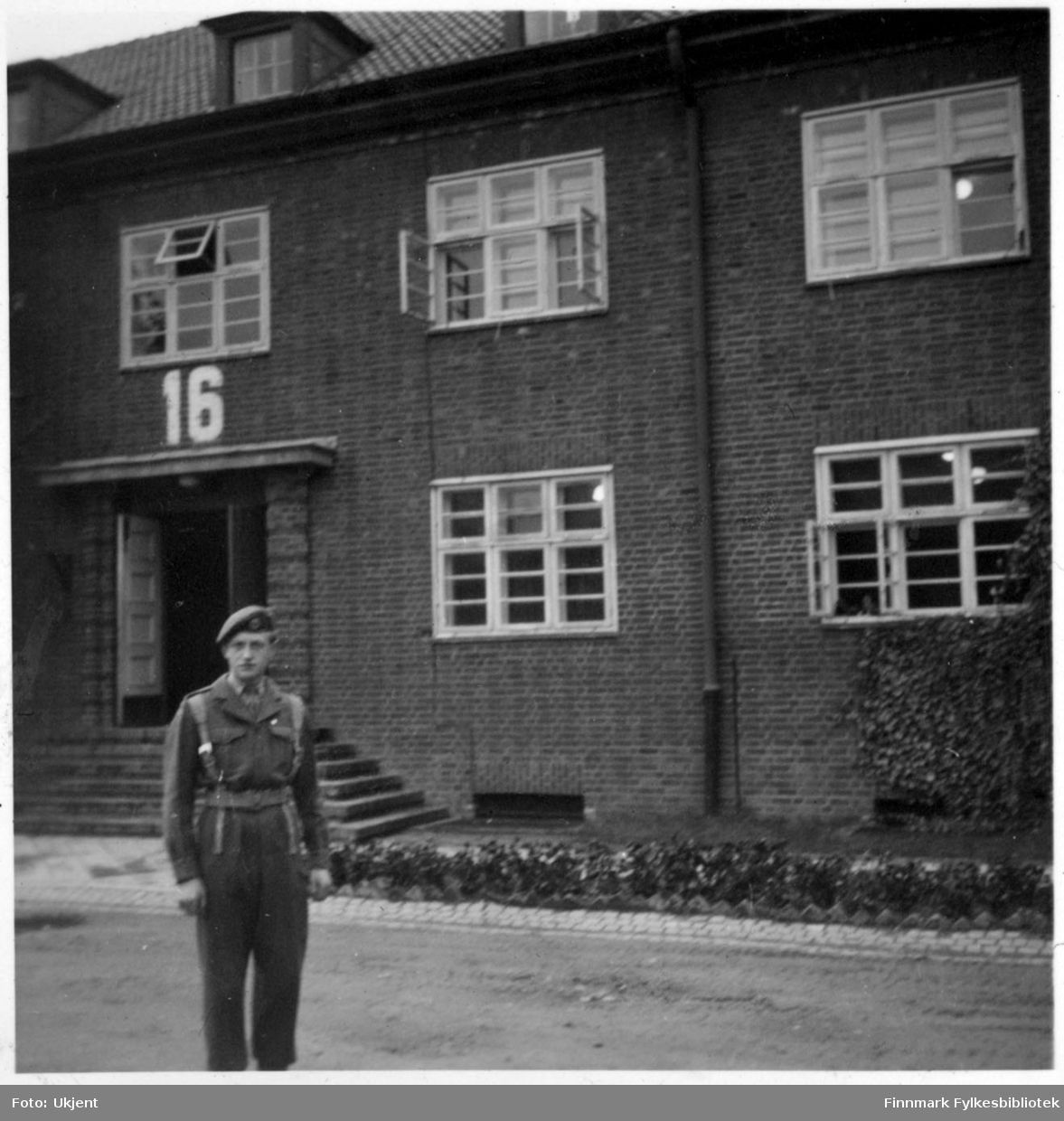 Reidar Martinsen i Tysklandsbrigaden etter frigjøringen.Han er kledd i uniform og står utenfor et murhus markert med nummeret '16'. På huset kan man se at enkelte av vinduene står åpne. En trapp fører til en åpen dør.