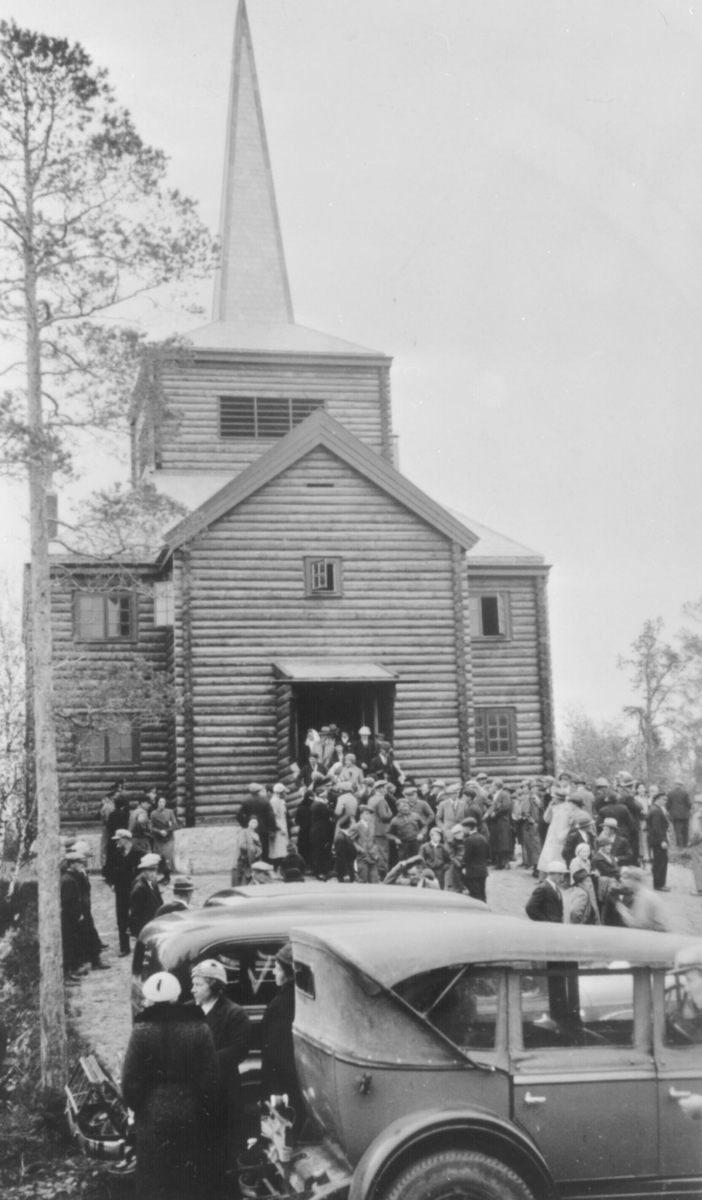 Gudstjeneste i Svanvik kapell er over. Folk er på vei ut av kapellet.Tre biler fra 1930-åra står i forgrunnen. Det er mye folk på bildet. Kapellet ble bygd i rundtømmer og ble innvidd i 1934.