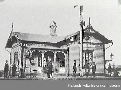 Veddige järnvägsstation som ny år 1880 utmed Varberg-Borås Järnväg, invigd samma år. Stationen är rikt utsmyckad med figursågade dekorer i tidstypisk eklektisk stil, dvs hämtar inspiration från och sammanför element från olika arkitekturstilar. Flera personer står på perrongen varav flera, kanske samtliga, förmodligen arbetar vid järnvägen och stationen.