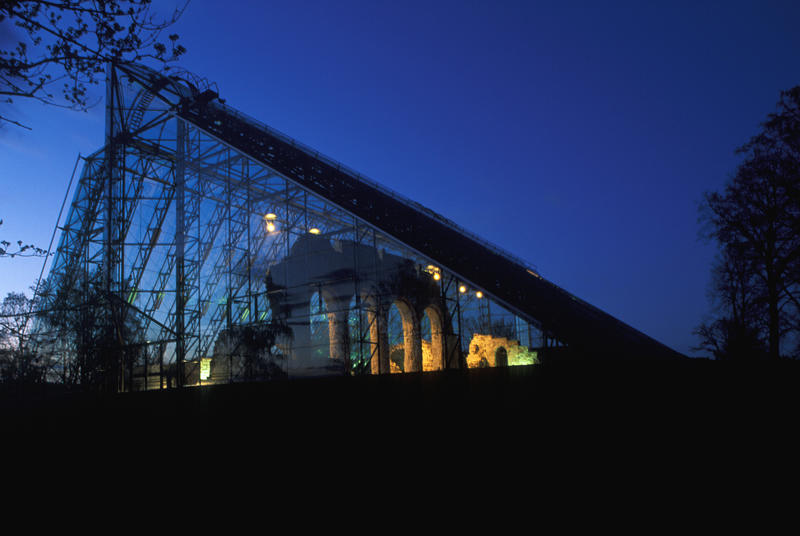 Domkirkeruinen under vernebygget i glass og stål er delvis opplyst og delvis i silhuett mot en mørkeblå kveldshimmel.