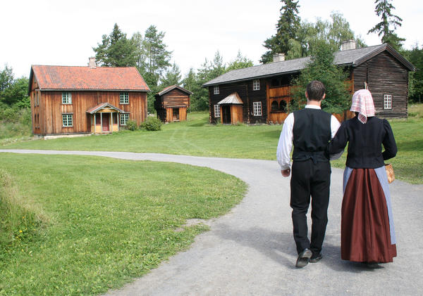 A couple dressed in traditional Norwegian costumes are walking arm in arm towards two old farm houses.