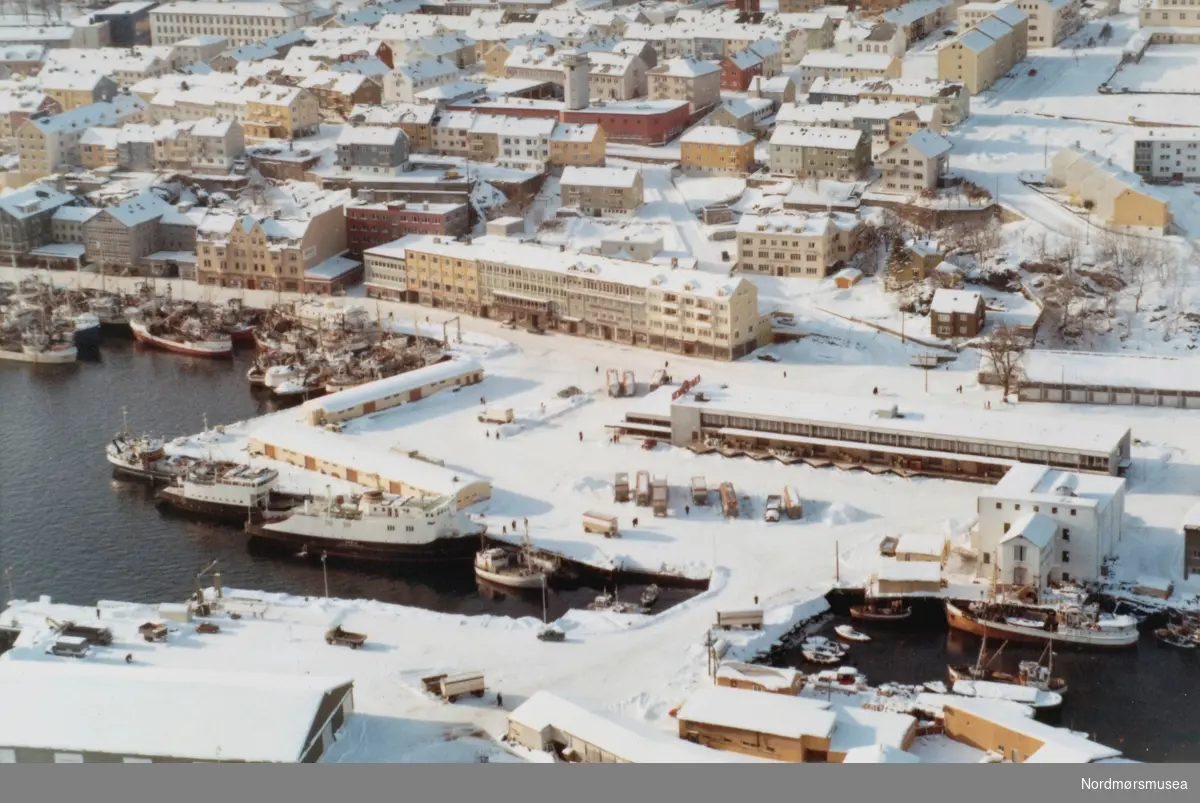 Flyfoto fra a/l Rutebilstasjonen på Nordmørskaia på Kirkelandet i Kristiansund, med Devoldholmen helt i front. Flyfotoet er utført av Widerøes flyveselskap den 23. februar 1969. Fra Nordmøre museums fotosamlinger. Reg: EFR
