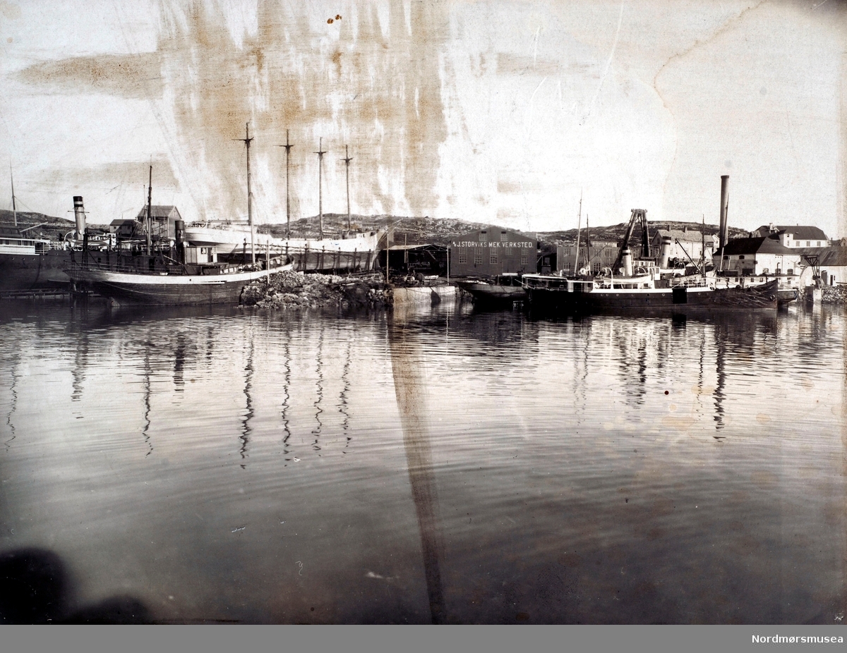Bilde av J. Storviks Mek. Verksted på Dahle ved Kristiansund, sett fra sjøen. Det er rekke båter ved kaier og på patentslippen.
Ved kjelkrankaia ses den første kjelkrana bygd av massivt tømmer i ca. 1903 og det ligger 2 rutedampskip, ytterst D/S”Kværnes (1)” tilhørende Kværnes og Grip Dampskibsselskab og innenfor ligger D/S”Hankø” og det ligger en fiskekutter innerst, som vi ser hekken på til venstre for de 2 rutebåtene foran kjelkrankaia. Dette er sansynligvis verkstedets fiskekutter som ble til D/S”Villa” bnr.10 til Fyrvesenet som ligger til utrustning. 
På den nye patentslippvogna klinkbygd i stål 1918, den største slippen mellom Bergen og Trondheim, står 2 båter, forrest et 4-mastret seilskip, et stort finsk skonnertskip S/S”Regina” som er til reparasjon etter grunnstøting og bakerst damprutebåten D/S”Smølen”  til Møre Fylkes Ruteselskap (MFR). Over ”Smølen” ses demningen på Nydammen (Storvikdammen).
Foran baugen på ”Regina” ses blikktaket over maskinparken, lokke-og klippemaskin, platevals, spantbukkemaskin, ved innkjørselen til verkstedet.
På nordsiden av piren foran bakerste båt på patentslippen ses damperen D/S”Bjugna”, tilhørende Julius Loennechen med flere, som får innsatt maskin og overbygg.
Piren er bare delvis utfyllt og bygd.
Over bakerste båt på patentslippen ses bolighuset til verkstedet med uthus, bygd ca. 1910 og brant på 1980-tallet, hvor familien Hjelmaas bodde, senere familien Paul Storvik. 
Bolighuset var et gammelt meieri i tømmer flyttet fra Straumsnesset og bygd opp ca. 1910 og brant på 1980-tallet.
Til høyre for Regina ses maskinverkstedbygningen med firmanavnet på veggen, bygget etter brannen i februar/mars 1911 og revet etter 1980. Over høyre del av taket på maskinverkstedbygningen ses mønet på bolighuset til John Olsen Storvik, senere barna hans og revet etter 1980. 
Til høyre for maskinverkstedsbygningen ses først sjauer og malerbygget deretter lagerutleveringsbygget, med kontorer for timeskriver og verksmester i 1. etasje, og foran lagerbygget ses den første kjelkrana av massive tømmerstokker fra 1903, den kraftigste krana i Kristiansundsområdet. Over lagerutleveringsbygget ses bolighuset til familien Nils Holtan, senere familien Bastian Holtan. 
Til høyre for kjelkrana ses bolighuset til familien Ole Storvik, senere Karen Storvik, senere familien Schamaun og mellom det og pipa på kobberslagerverkstedbygningen ses, fra venstre, bolighuset til familien Nils Todalshaug, senere Anders Todalshaug og bolighuset til familien Otto Knudsen. Over taket på kobberslagerverkstedbygningen ses ”Murgården”, verkstedets flerleilighetshus for ansatte, bygget ca. 1910 og revet 1981. Til høyre for ”Murgården” ses mønet på bolighuset til familien Kristian Stabben senere familien Otto Kvendseth. Til høyre for baugen på ytterste båt, ved kjelkranakaia, ses en mekanisk svingbar kaikran, som bl.a. ble brukt til å losse ved til vedlageret i påbygget på høyre side av kobberslagerverkstedbygget og helt til høyre kan ses et kombinert sag- og tremateriallagerhus. Man kan også skimte et trematerialbygg, bak og mellom kobberslagerverkstedbygget og lagerutleveringsbygget ved grensegjerdet til verkstedet. Utenfor gjerdet gikk veien ned til sundbåttillegget på Dahle som lå like utenfor bildekanten.
På dette tidspunkt var verkstedet blitt et av de mest praktiske, velordnede og tidsmessige verksteder i sitt slag i landet.
Bildet er tatt sommeren 1920.
Samme bilde som FAKf-100296.220422 / KMb-1982-010.0157.
Bildet er på forsiden av prospektkortet FAKf-100296.220423 / KMb-1982-010.0158.jpg  (Info: Peter Storvik) ---
Storvik Mekaniske Verksted sett fra sjøsiden. Det er flere fartøyer på slipp, samt flere dampbåter fortøyd ved kaien. Her ser vi også den finske seilskuten ";Regina"; som er til reparasjon etter en grunnstøting. Dampbåten på slipp er ";Jotunheim";, mens båt ved kao er ";Kvernes";. Fra slutten av 1800-tallet og frem til midt på 1900-tallet var Storvik Mek. Verksted AS og Sterkoder Mek. Verksted to hjørnesteinsbedrifter i Kristiansund som bygde skipsmotorer, reparerte fartøy og bygde mindre båter. Det var mange opp- og nedturer da som nå. I 1966 gikk Sterkoder Mek. Verksted over til mer ensidig å bygge båter (Kamsvågperioden). Selskapet tok over Storvik Mek. Verksted i 1982. Sterkoder Mek. Verksted var i fremste rekke når det gjaldt effektiv skipsproduksjon, og det var under perioden med blant annet serieproduksjon av trålere at industri- og lagerhallene på Dale Industripark ble bygget og modernisert. Leveranser til olje- og gassvirksomheten ble enda et bein å stå på for Sterkoder Mek. Verksted, og anlegget i Smedvågen ble bygget opp til formålet. I febr. 1991 ble Sterkoder Mek. Verksted overtatt av Umoe-konsernet og er i dag en del av Umoe Eiendom etter at skipsbyggingen ble avviklet rundt 2003. Umoe Sterkoder AS sine anlegg består i dag av industriområdet i Melkvika og Dale Industripark på Dale. Industriområdet i Smedvågen ble solgt i des. 2006. Aibel AS leier området i Melkvika i sin helhet. Dale Industripark omfatter Umoe Sterkoder sitt anlegg på Dale og leies ut til flere ulike aktører. Umoe Sterkoder AS og leietakerne er hver for seg solide leverandører til offshore-industrien innenfor sitt felt. Alle har forskjellige tjenestetilbud som gjør at de samlet fremstår som bra. Fotograf er J. O. Engvig. Datering er ukjent, men trolig omkring 1900-1910. Fra Nordmøre Museums fotosamlinger.