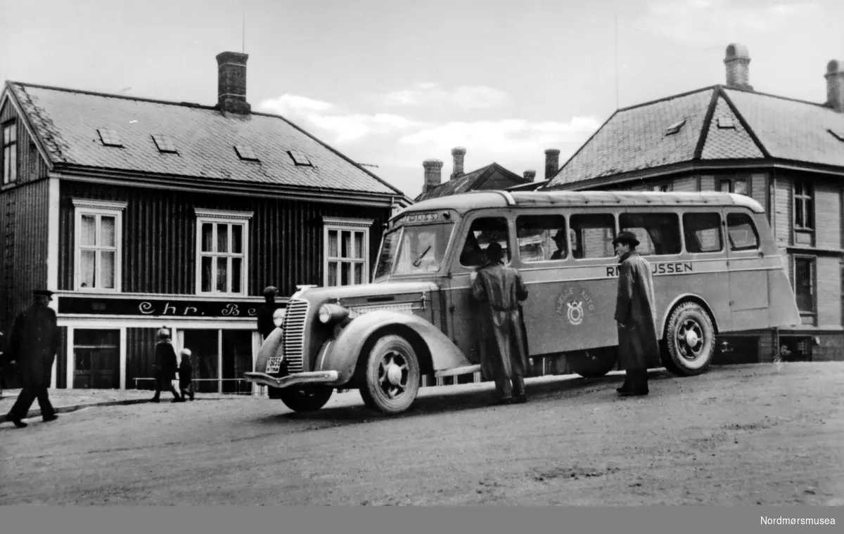 Postkort: ";Ringbussen, Kristiansund N. Eneret G.
Sverdrup A/S 1937, 201";. Bussen fotografert på Torget på Kirkelandet i Kristiansund i 1937. Ringbussen er fra ";Hjelle Auto"; og har registreringsnummeret T-5056. Buss: Diamond T 1936-37.

Historien om Ringbussen i Kristiansund er lite kjent. Ruta ble etablert i 1936 og gikk inn i KFB i 1941. KFB ble etablert i 1934 under navnet Frei Billag, men rutene til Kr-sund kom først i gang i 1940 etter åpningen av Omsundbrua. Sveinung Berild skev høsten 2014 en artikkel  om Kristiansund Freil Billag (KBF) i bladet ";Rutebil";.

I bakgrunnen ser vi Chr. Berseth manufakturforretning til venstre og Grefsnes glassmagasin til høyre. Ellers så ser vi to menn som slår av en prat med bussjåføren, samt flere andre i bakgrunnen. Prospektkortet er fotografert og utgitt ved Georg Sverdrups Bokhandel i 1937. Forretninger, butikker, mennesker, menn, barn. (Informant: Sveinung Berild og Nordmøre Museum). Fra Nordmøre Museums fotosamlinger.
