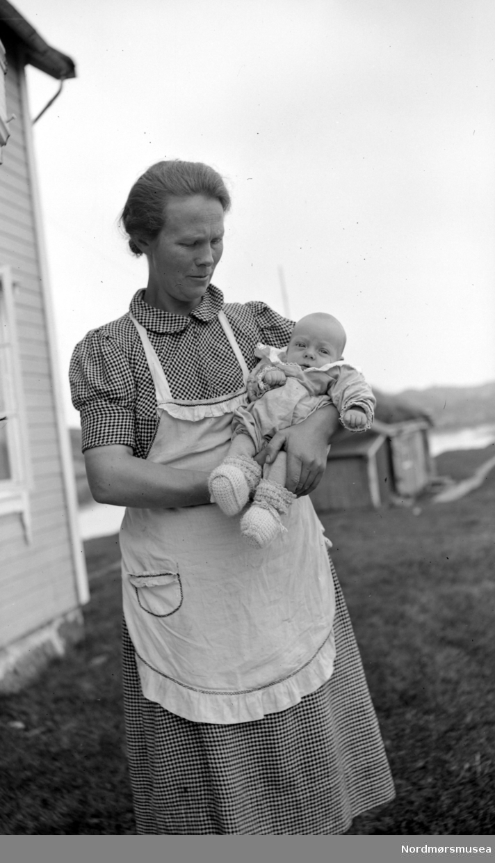 Foto av en kvinne med et barn på armen. Fotografert trolig på Langøya på Vevang i Hustadvika kommune. Smørholmen i bakgrunnen. Fra Nordmøre Museum si fotosamling