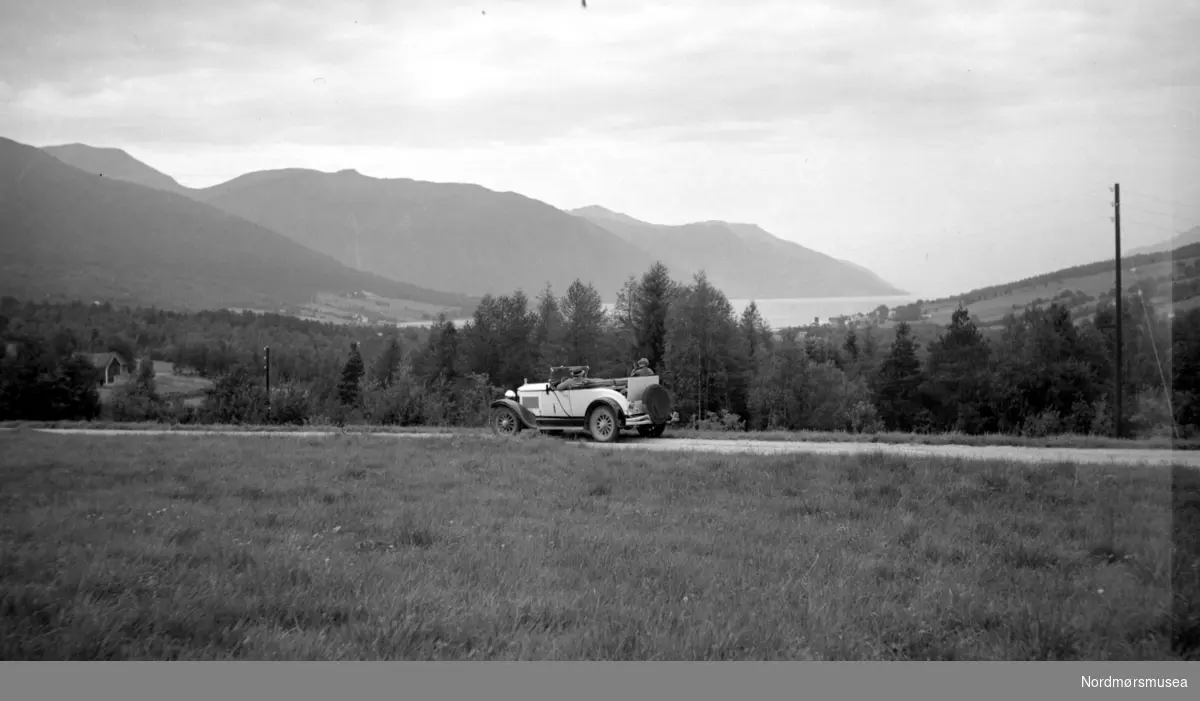 Er vel muligens på Tiltereidet, mot Eidsvåg og Langfjorden. (E Sæbø) . - 
bil langs en landevei. i Møre og Romsdal fylke. fjell, fjord, stolpe, eng. Datering er ukjent, men sannsynligvis omkring 1920 til 1939. Fra Nordmøre museums fotosamlinger.
