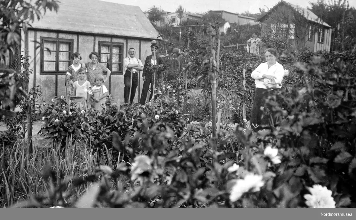 Foto trolig fra en kolonihage. To menn, fire kvinner og to barn poserer for fotografen foran en liten hytte. Kan det være Minde havekoloni i byskogen i Kristiansund? Det er ukjent hvem vi ser på bildet. Datering er også usikkert, men trolig omkring 1930 til 1939. Fra Nordmøre museums fotosamlinger. /Reg:EFR2013/
