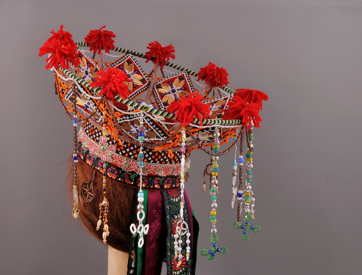 Brudekrone, nylaga.
"Pull" truleg med botn av stiv kartong og kledd med kraftig raudt klede. Påsydd (frå nedkant og oppover) eit band med satengbotn lansert blondemønster og påsydd kvite glasperler, ein sylvknipling og eit perlebendel. Perlebendelet er tredd med glasperler i svart, kvitt og orange.Sjølve krona er oppbygd rundt eit stativ av stål, raudlakkert. Heile stativet er dekt av glasperler i ulike fargar. Inni kvart "rom" er det eit sydd raudt kvadratisk klede som er pryda med glas- og stavperler av åttebladrose-mønster på begge sider, alle kvadrata er like og dei er montert på høgkant. I tillegg er det ein del ulike heng frå øvre kant av krona, her er det nytta ulike typer glasperler. På toppen av krona er det festa små duskar av laustvinna, raudt ullgarn i kvart skjæringspunkt. Inni krona, over hovudet, ligg eit ubleika band i kryss og i krysset er det sydd fast ein rosett.
Bak på krona er det sju heng av ulike band. På kvar side er det "øyrepynt". Frå sida og bakover er det 11 heng med ulike typer perler.