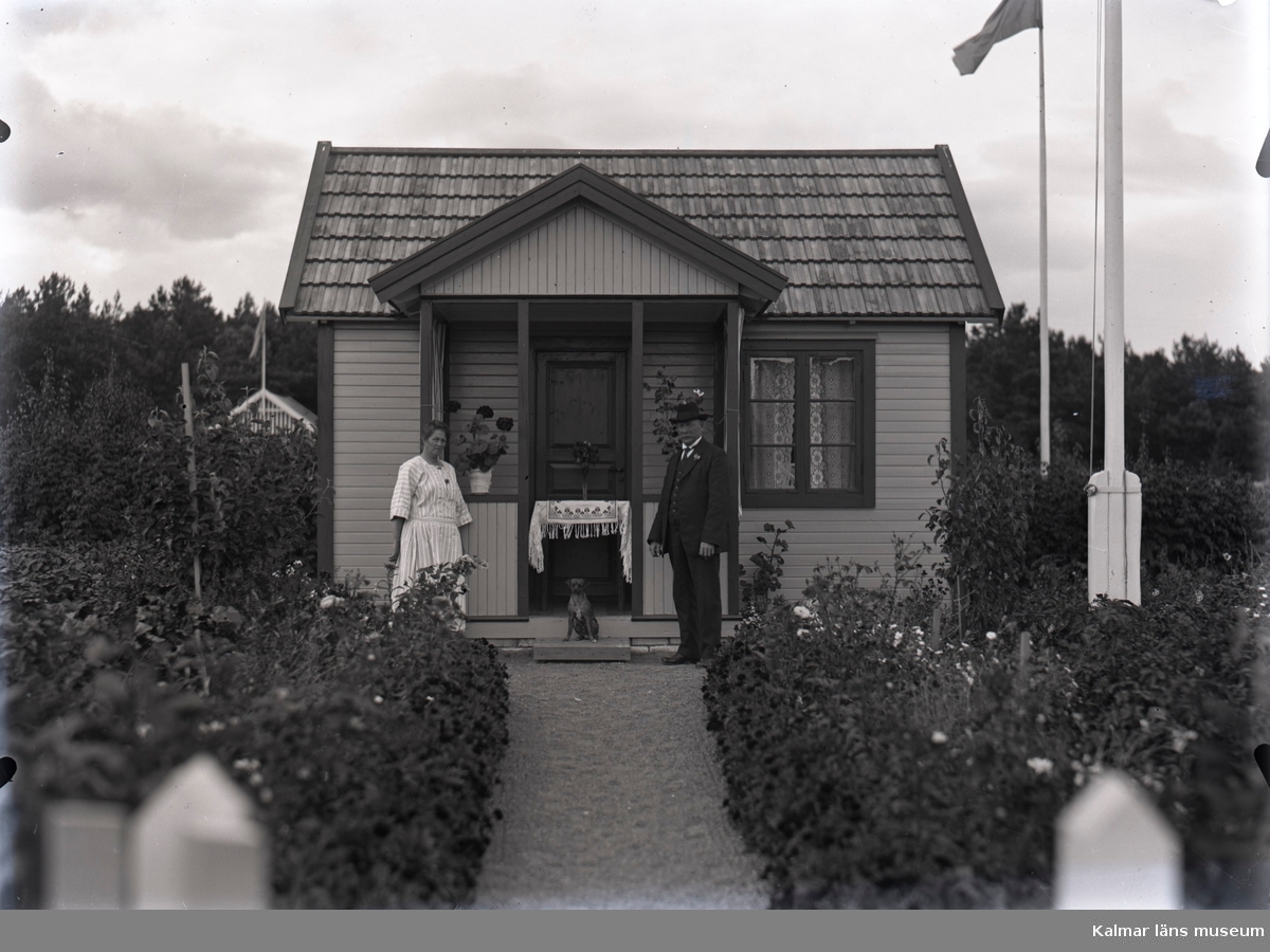 En stuga, man, kvinna och hund i Kalmar södra koloniområde, fotograferat omkring 1930. Kalmar södra koloniförening grundades 1917 och har idag 105 kolonilotter. Området ligger strax söder om länssjukhuset i Kalmar med huvudingång från Stensbergsvägen.